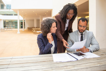 Business team of three studying documents in street cafe. Business man and women sitting and standing at table outside and staring at documents. Paper expertise concept