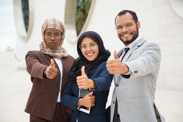 Fototapeta na wymiar Multicultural business group posing and making like gesture. Businessman and Muslim businesswomen standing outdoors, showing thumb up and smiling at camera. Successful business team concept
