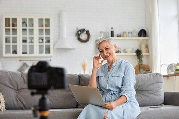 Successful senior female blogger sitting on sofa with laptop in front of camera on stand, filming...