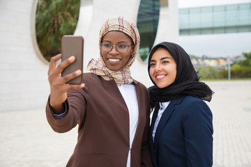 Happy female office friends taking selfie outside. Muslim business women in hijabs standing outdoors, holding mobile phone, posing and smiling at screen. Photo concept