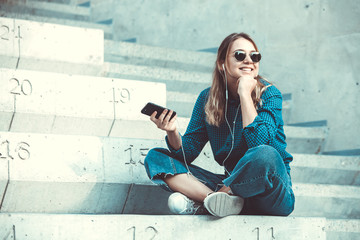 Portrait of a happy girl listening music on line with headphones from a smartphone in the street in a summer sunny day. Woman listening to music with the phone and having fun