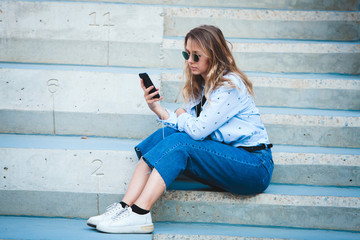 Portrait of a happy girl listening music on line with headphones from a smartphone in the street in a summer sunny day. Woman listening to music with the phone and having fun