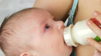 A two-month newborn baby lies on the mother arms and sucks a nipple from a bottle of milk.