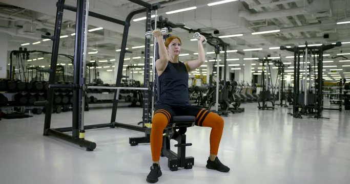 sporty woman is tensing muscles of her hands, lifting dumbbells, sitting on training equipment