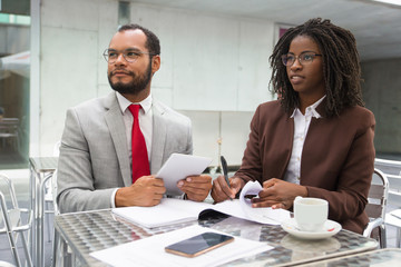 Business colleagues reviewing documents in street coffee shop and staring away. Business man and woman sitting in cafe, checking papers, holding tablet and looking away. Meeting in cafe concept