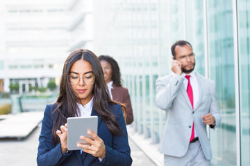 Obraz premium Confident businesswoman using tablet while going down business street. Business people with gadgets walking behind her. Wireless technology concept