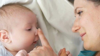 Young beautiful mother with long dark hair is lying on a white bed and playing with a newborn baby of two months