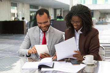 Cheerful business colleagues checking documents during coffee break. Business man and woman sitting in cafe, reading documents and using tablet. Paperwork concept