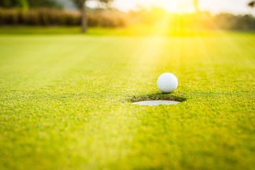 A white golf ball on green grass near hole with golf course background,green tree sun rays. 