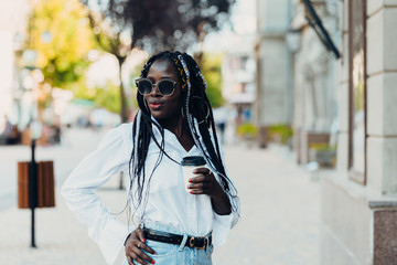 Portrait of a smiling young dark-skinned girl with pigtails in glasses with coffee in hand outdoors on a sunny day.