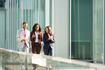 Business team moving along outdoor office glass wall. Business man and women walking outside, using tablet, talking, smiling, laughing. Discussion outdoors concept
