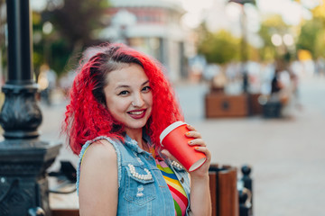 Stylish happy young woman in a bright striped sundress and a denim T-shirt with red hair walks along the street. Portrait of a smiling girl with a red paper glass of coffee