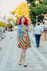 Stylish happy young woman in a bright striped sundress and a denim vest with red hair walks on the street. Portrait of a smiling girl