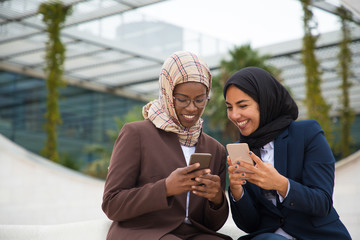 Happy office colleagues chatting on smartphones and sharing news. Muslim business women in hijabs sitting outside, using mobile phones together, texting messages. Communication concept