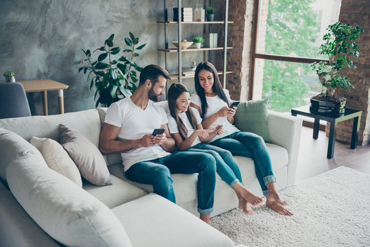 Nice attractive lovely cheerful cheery friendly adorable healthy family wearing casual white t-shirts jeans sitting on sofa using 5g app browsing at industrial loft style interior living-room