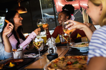 Group of young female friends having fun in restaurant, toasting with wine talking and laughing while dining at table.