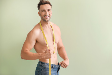 Handsome muscular man in loose jeans and with measuring tape on color background. Weight loss...
