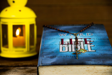 Elegant holy bible and Christian crucifix with rosary beads with light candle in yellow lantern on wooden background.