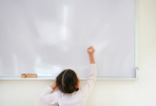 Little Caucasian Child Girl Writing Something On Whiteboard With A Marker In The Classroom. White Board With Copy Space For Text. Back View.