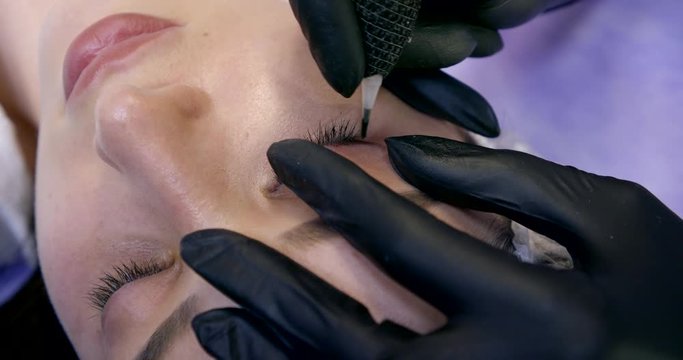 master is making tattoo of arrows on eyelid of female client, closeup view of hands