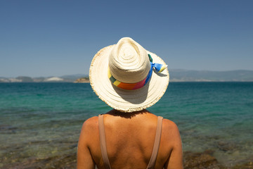 Fototapeta na wymiar Back view of young woman in white hat standing towards the sea.