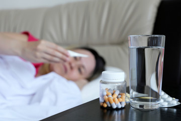 Sick woman lies in a bed with digital thermometer in hand, cold and flu season. Pills and water glass in the foreground, concept of illness, fever, high body temperature