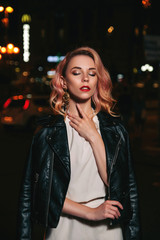 Luxury beautiful blond woman with curly hair and evening make up standing in the middle of night city. She wears casual white blouse and trousers and leather jacket