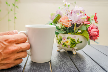 A man's hand holds a white Cup of coffee on a wooden table close-up, next to a bouquet of flowers