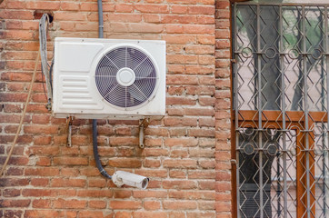 White air conditioning on the brick wall of the building near the window with bars, under the air conditioning is a surveillance camera
