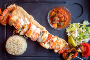 Roasted pieces of white and red fish fillet on a skewer with garnish, sauce and salad. Side dish of rice and stewed vegetables, served fish dish, top view, Mediterranean style