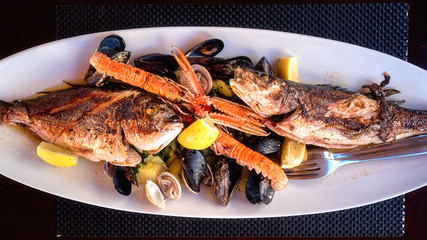 Traditional Mediterranean grilled seafood dish, plate with fish, shrimp, mussels, lemon and boiled potato, served food, top view