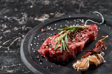raw meat steak with rosemary twig sprinkled with salt and pepper near garlic and dried cayenne on...