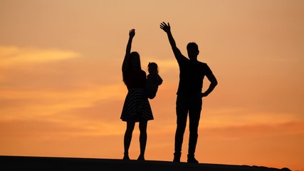 Young family with little baby silhouetted against sunset sky, wave hand a farewell to camera. Mother hold infant on hands, father stay near. Vivid orange colours of summer sunset on background