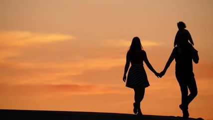 Young family of three walk against sunset, man carry tiny child on shoulders hold woman by hand. Silhouetted slow motion shot, beautiful colours of clouds at summer evening seen on background