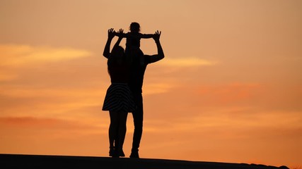 Cheerful young family silhouetted shot against yellow sunset sky. Happy people look to camera, woman jump and dance, man rise up baby hands. Positive mood, summer evening time