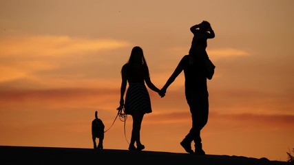 Young family of three walk together, black silhouettes against yellow sunset sky. Dad carry little daughter on shoulders, holding woman by hand. Dog run near. Calm and warm summer evening time