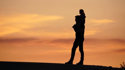 Father stay with little baby on shoulders, shadow figure against vivid yellow sunset sky. Man step and turn, standing still then