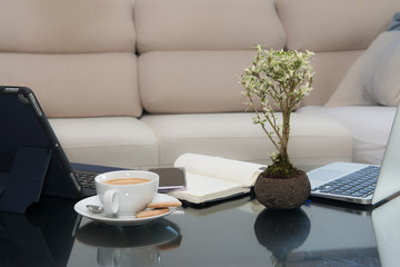 Glass table with a tablet, a cell phones notebook, a cup of coffee and a plant and in the background an elegant cream armchair