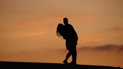 Happy couple at top, man embrace lovely woman and lift her up, girls legs fly in air. Silhouetted shot against sunset sky. Life is good concept
