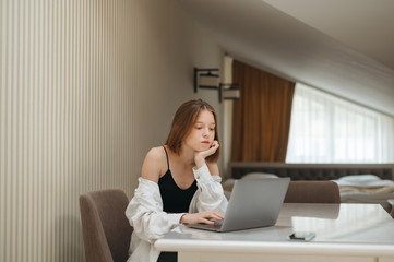 Attractive girl sits at a table at home and uses a laptop, looks intently at the screen, wears stylish casual clothes. Student studies in his room at the table, uses a laptop.
