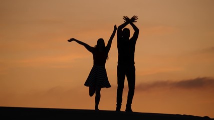 Cheerful couple wave hands and jump up, silhouetted full length shot against sunset sky. Two people stay on top and actively move arms, try to take attention, looking to camera
