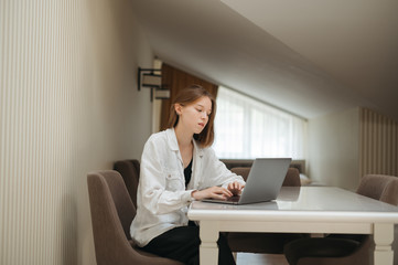 Beautiful girl student in casual clothes sits in the apartment at the desk and works at a laptop. Attractive young female freelancer working at home at computer.