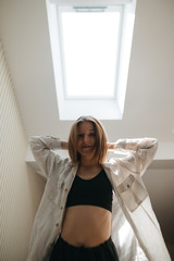 Happy girl in a black bra and shirt stands against a light attic window, looks into the camera, straightens her hair and smiles. Silhouette portrait in the apartment