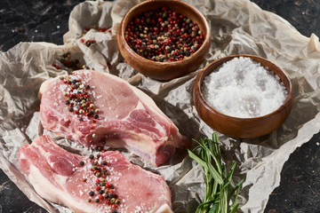 raw pork steaks on parchment paper near wooden bowls with salt and pepper on black marble surface
