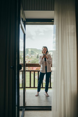 Full length portrait of a girl standing on a balcony in an apartment with a smartphone in hands,having fun, wearing stylish casual clothes.Stylish woman stands on a balcony with a beautiful landscape