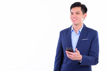 Happy young handsome Asian businessman thinking while using phone