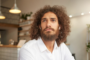 Closeup portrait of attractive curly guy with lush beard posing over cafe interior, wearing headphones and white shirt, looking aside pensively, waiting for his order