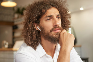 Young beautiful dark haired man with beard posing over coffee house, looking in front of him with pensive and concentrated look, holding his chin