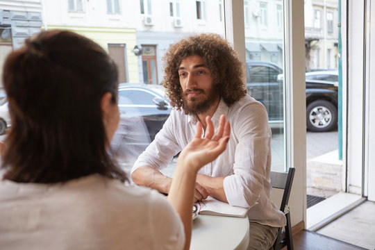 Indoor portrait of beautiful curly male with beard having meeting in cafe, looking attentively and calmly on female next to him, sitting at table by the window