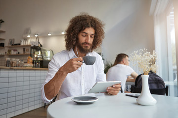 Fototapeta na wymiar Young attractive male with lush beard and brown curly hair watching videos on his tablet while having cup of tea, posing over cafe interior